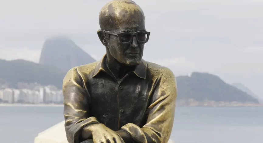 Estátua de Carlos Drummond de Andrade na Praia de Copacabana. O poeta está sentado num banco da orla.